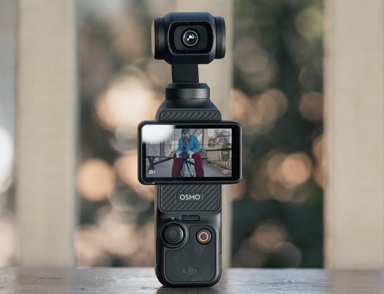 The camera standing on a table, screen on, with a blurred background behind it